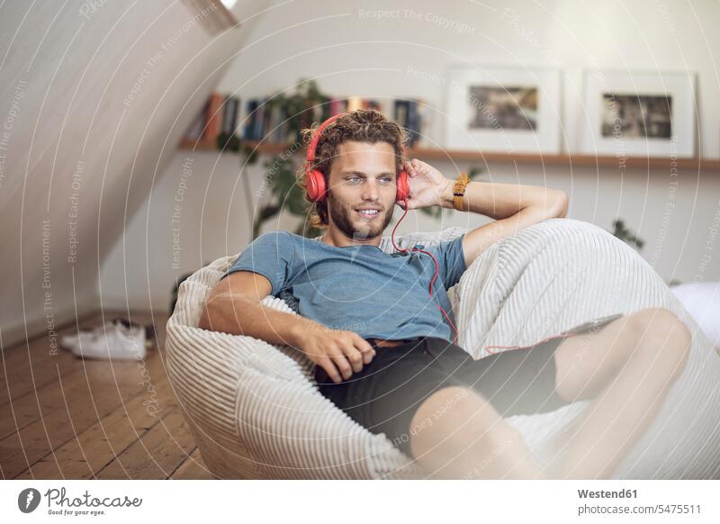 Young man relaxing in beanbag at home listening to music students human human being human beings humans person persons curl curled curls curly hair T- Shirt