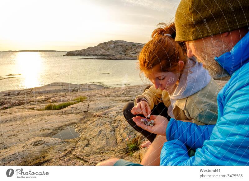 Sweden, Vastra Gotaland County, Grebbestad, Man and teenage girl collecting pebbles on rocky shore of Tjurpannans Nature Preserve at sunset outdoors