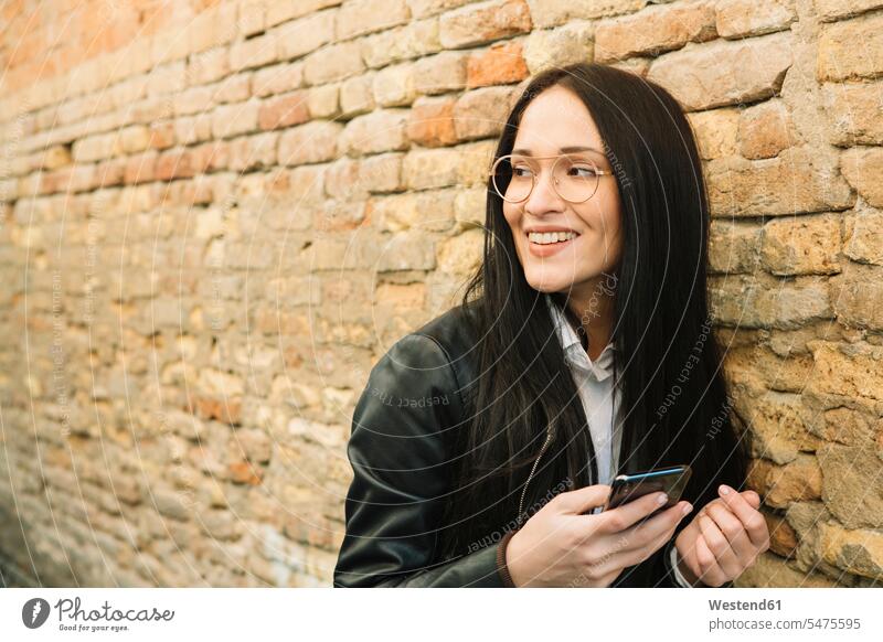 Smiling young woman with cell phone at brick wall looking around looking round look round look around mobile phone mobiles mobile phones Cellphone cell phones