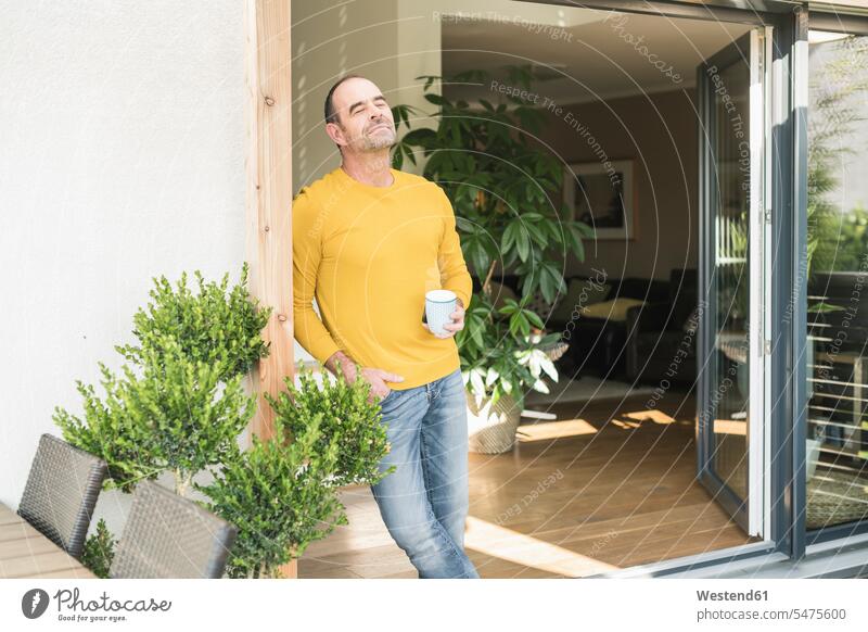 Mature man with closed eyes standing at terrace door at home with coffee cup human human being human beings humans person persons celibate celibates singles