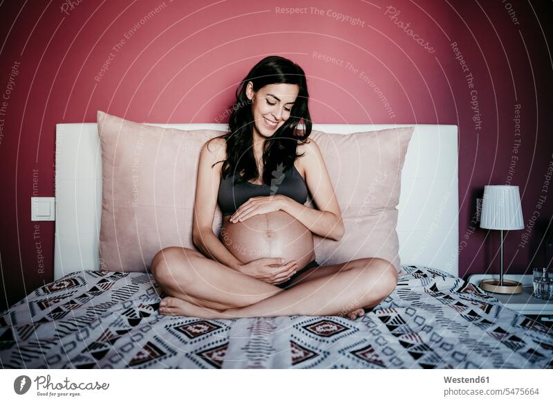 Happy pregnant woman looking at stomach while siting in bedroom at home color image colour image indoors indoor shot indoor shots interior interior view