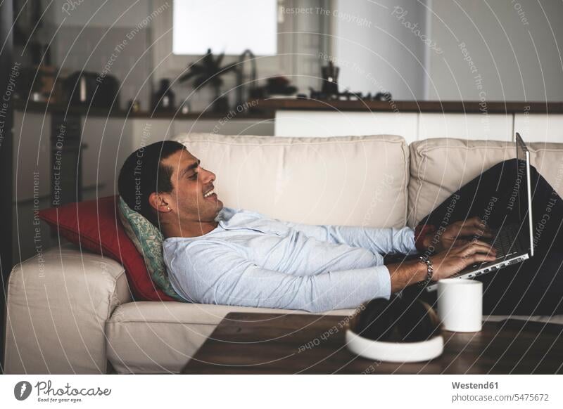 Smiling young man lying on sofa at home using laptop men males smiling smile laying down lie lying down couch settee sofas couches settees Laptop Computers