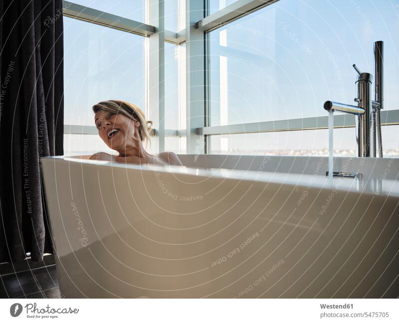 Cheerful blond senior woman taking bath in bathtub against window at luxury hotel room color image colour image indoors indoor shot indoor shots interior