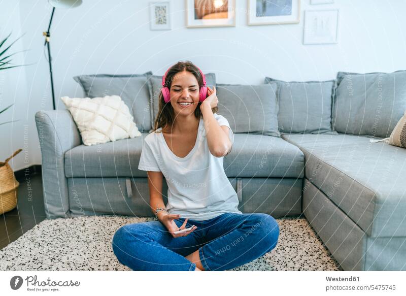 Happy young woman sitting on floor in living room listening to music with cell phone human human being human beings humans person persons caucasian appearance