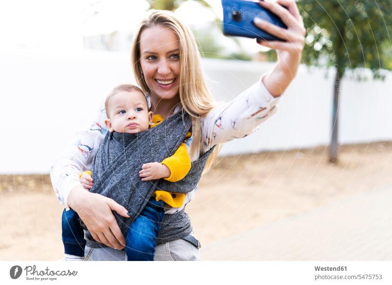 Happy mother with baby boy in a sling taking a selfie telecommunication phones telephone telephones cell phone cell phones Cellphone mobile mobile phones