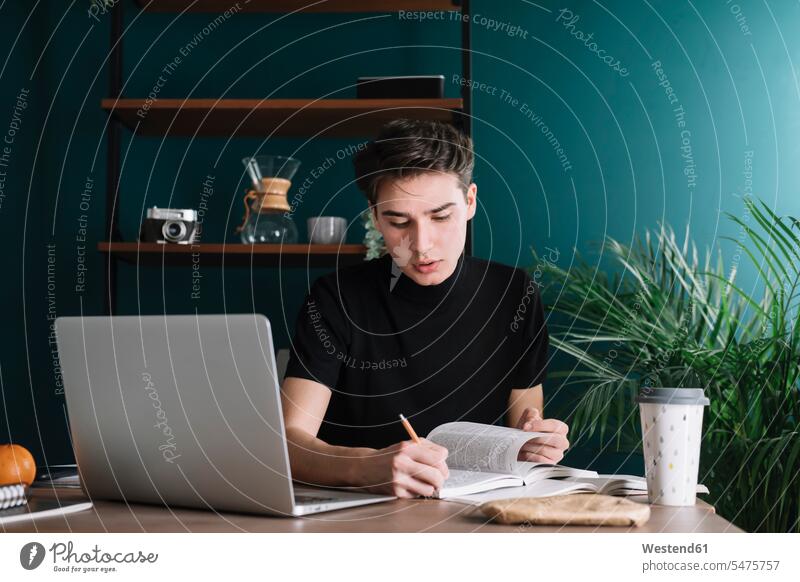 Male student writing in book while sitting with laptop at table doing homework color image colour image indoors indoor shot indoor shots interior interior view