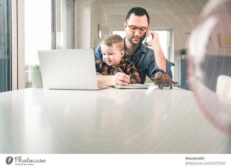 Busy father working at table in home office with son sitting on his lap Seated sons manchild manchildren At Work pa fathers daddy dads papa working from home