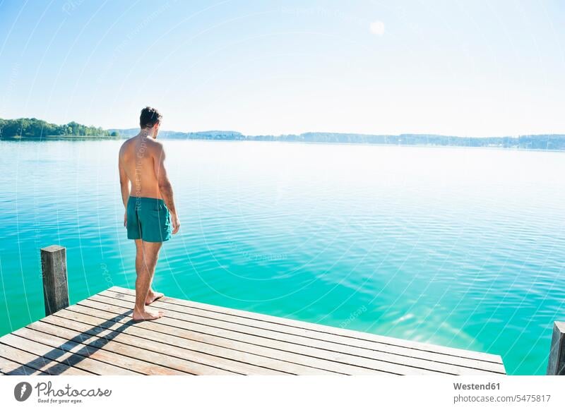 Back view of young man in swimming shorts standing on jetty looking at lake relax relaxing seasons summer time summertime summery free time leisure time