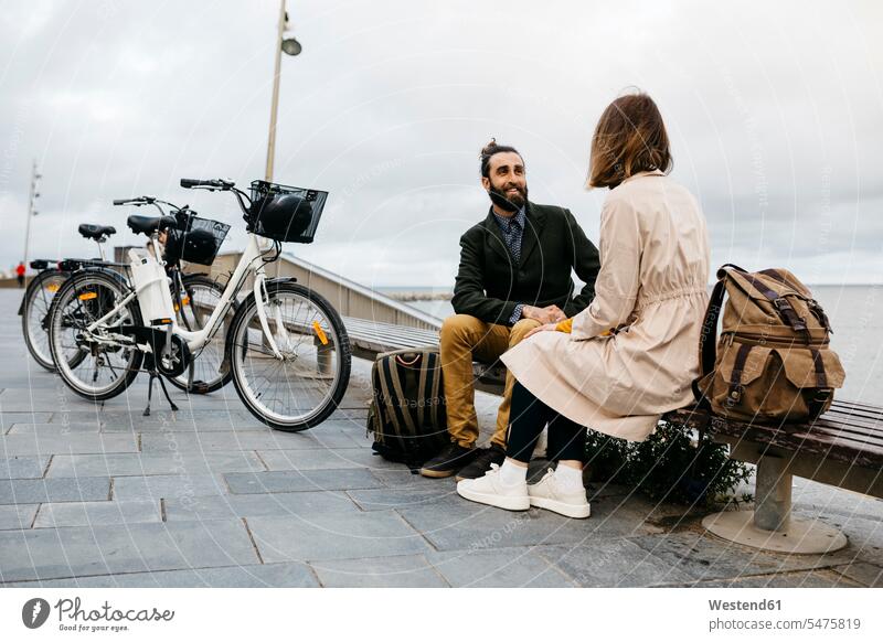 Couple sitting on a bench at beach promenade next to e-bikes talking sea front boardwalk E-Bike Electric bicycle Electric Bike speaking Seated couple twosomes