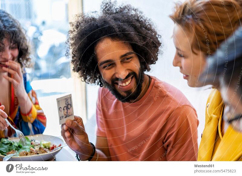 Happy man showing instant photo to friends having lunch in a restaurant dish dishes Plates images picture pictures photographs photos instant photograph