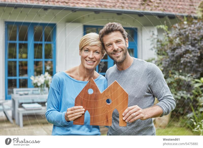 Portrait of smiling couple standing in front of their home holding house model models portrait portraits houses twosomes partnership couples smile building