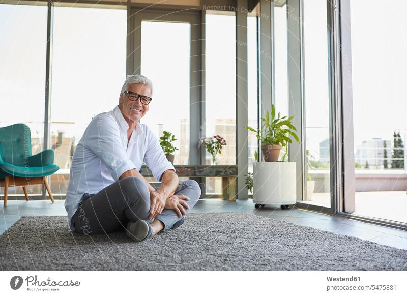 Smiling mature man relaxing sitting on carpet at home men males relaxed relaxation Seated smiling smile carpets rug rugs Adults grown-ups grownups adult people