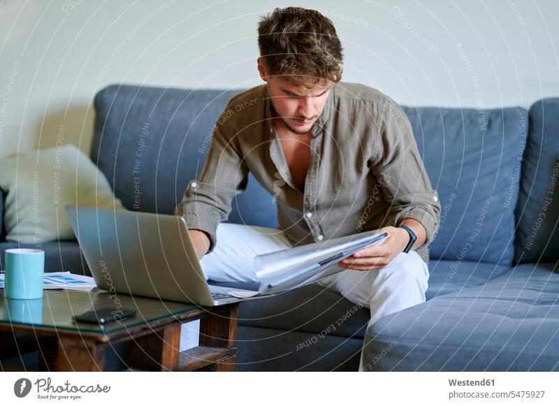 Young male freelancer reading documents while sitting on sofa with laptop at home color image colour image indoors indoor shot indoor shots interior