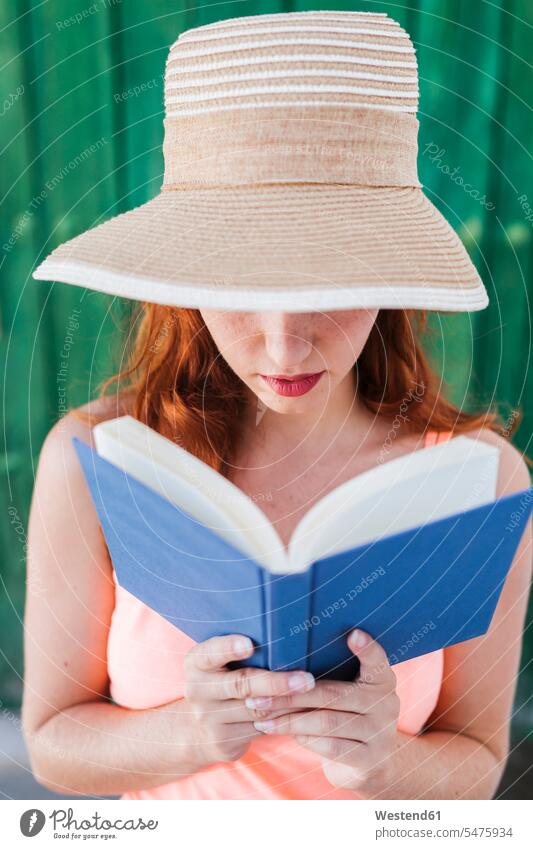 Redheaded young woman in front of green wooden door reading a book in summer books summer time summertime summery free time leisure time fashionable