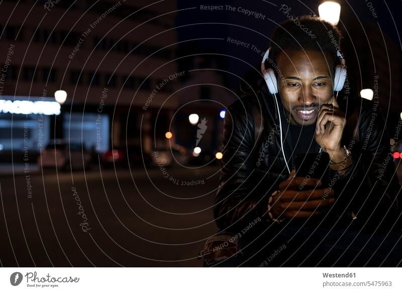 Man in the city at night with smartphone and headphones coat coats jackets headset telecommunication telephone telephones cell phone cell phones Cellphone