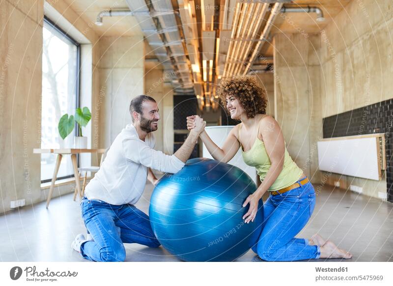 Happy man and woman arm wrestling on fitness ball in modern office kneeling Kneeing Fun having fun funny Competition competitive freelancer freelancing