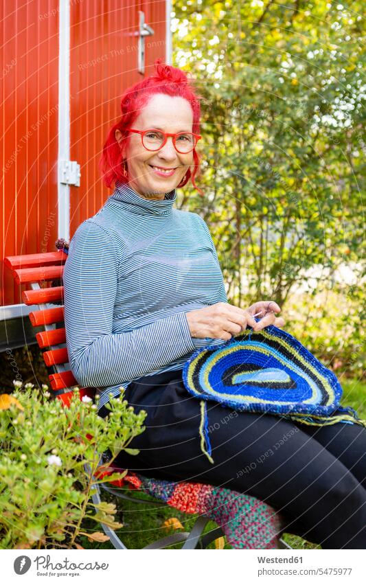 Portrait of smiling senior woman with red dyed hair sitting in front of red trailer in the garden crocheting portrait portraits coloured females women