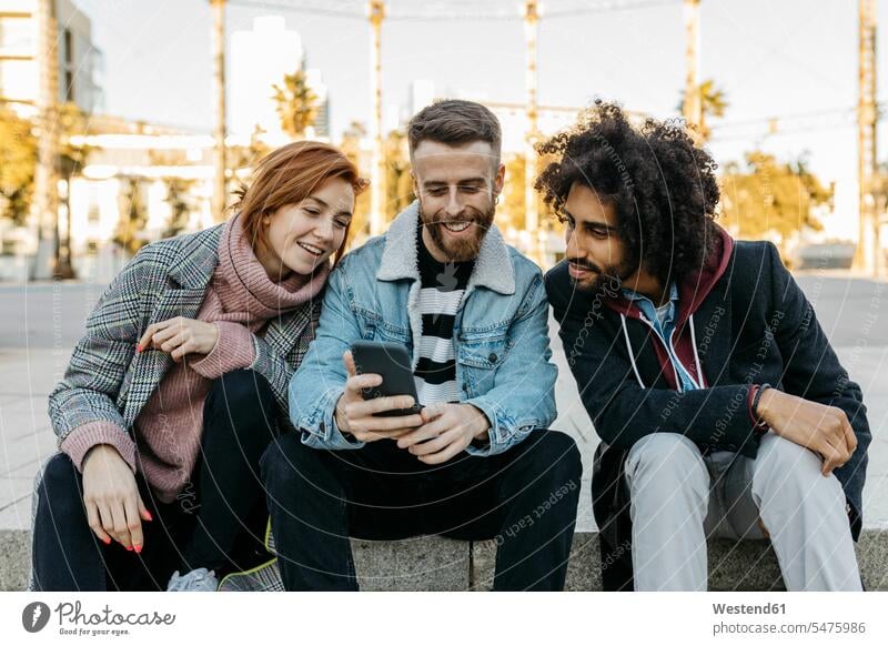 Three happy friends sitting outdoors looking at cell phone happiness Seated eyeing mobile phone mobiles mobile phones Cellphone cell phones view seeing viewing