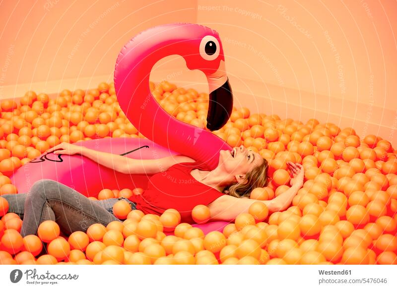 Cheerful young woman with inflatable flamingo lying in orange ball pit color image colour image leisure activity leisure activities free time leisure time