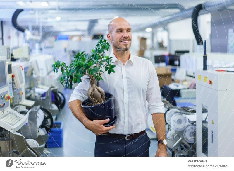 Smiling businessman holding potted plant while looking away at factory color image colour image indoors indoor shot indoor shots interior interior view