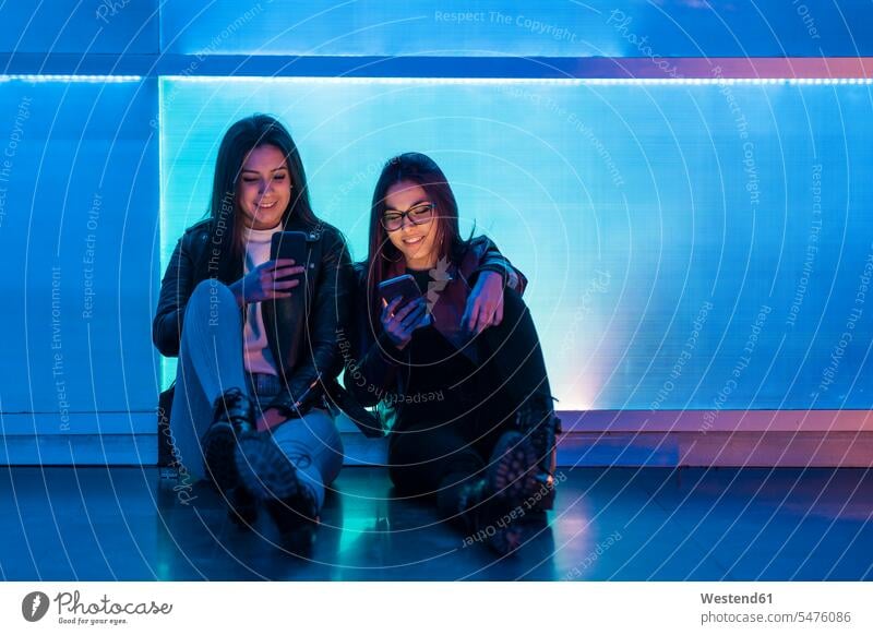 Portrait of two smiling teenage girls sitting in front of blue glass pane looking at smartphones human human being human beings humans person persons