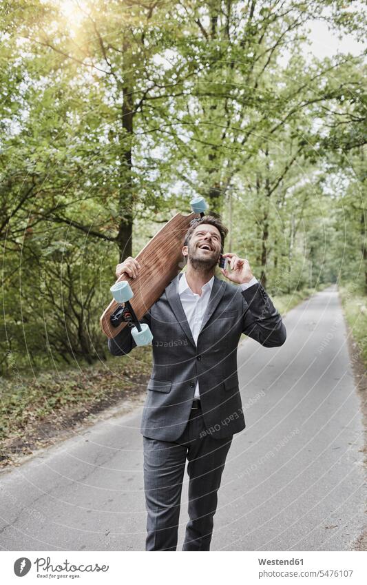 Happy businessman with skateboard talking on smartphone on rural road Skate Board skateboards Businessman Business man Businessmen Business men country road