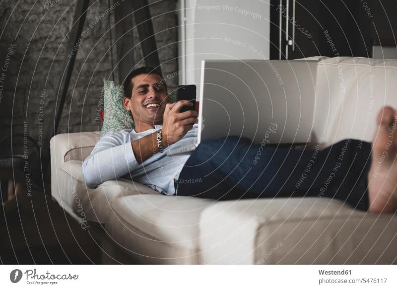 Happy young man lying on sofa at home using cell phone and laptop laying down lie lying down smiling smile men males Laptop Computers laptops notebook