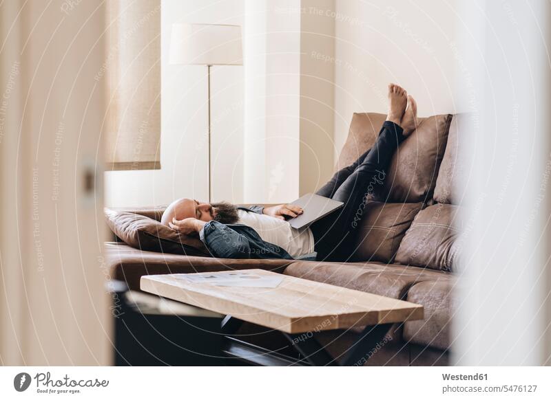 Man taking nap while lying with laptop on sofa at home color image colour image casual clothing casual wear leisure wear casual clothes Casual Attire