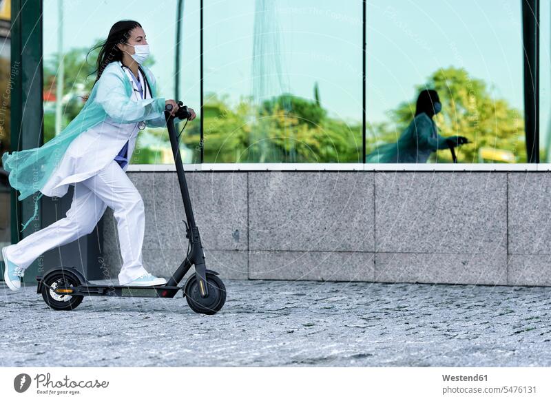 Young doctor in face mask and uniform riding push scooter while going to hospital during COVID-19 color image colour image outdoors location shots outdoor shot