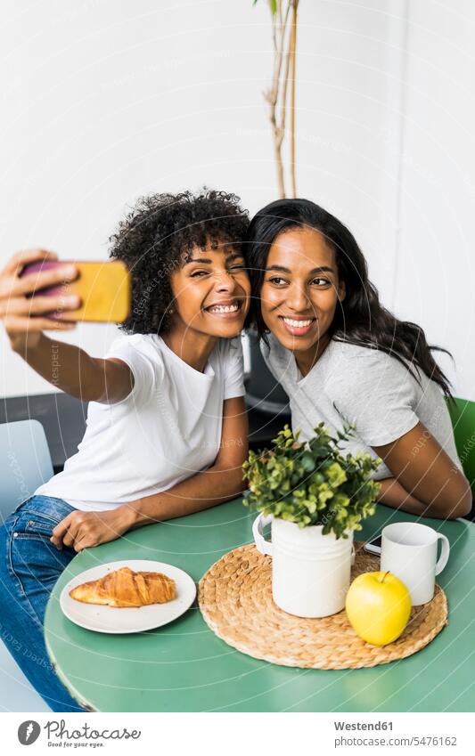 Two happy girlfriends sitting at table taking a selfie Seated happiness woman females women home at home together Table Tables Selfie Selfies female friends