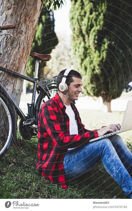 Young man sitting in a park wearing headphones and using laptop next to bicycle men males Laptop Computers laptops notebook bikes bicycles parks headset Seated
