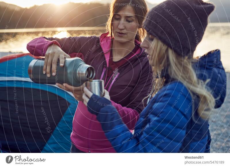 Two women with hot drink camping at lakeshore female friends Lakeshore Lake Shore lakeside tent tents woman females hot beverage hot beverages mate friendship