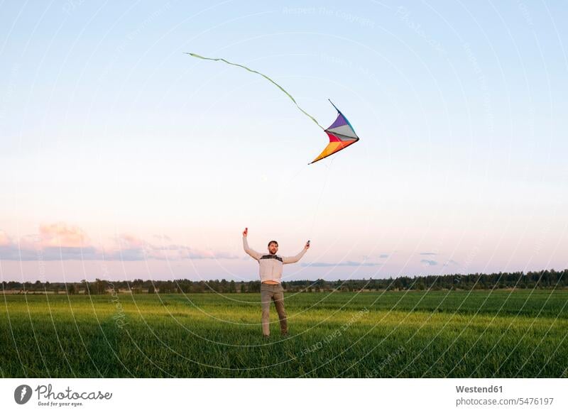 Mid adult man flying kite while standing on grassy landscape against sky at sunset color image colour image leisure activity leisure activities free time