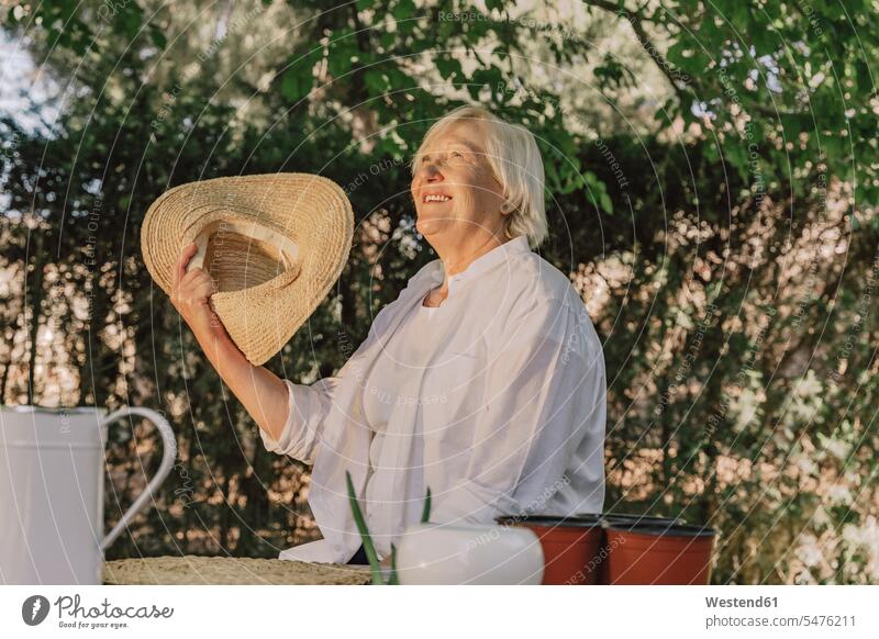 Cheerful senior woman holding hat looking up while sitting against tree in yard color image colour image Spain leisure activity leisure activities free time