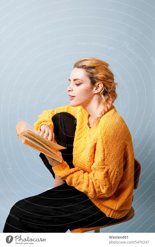 Young woman wearing yellow knit pullover in front of blue background reading a book books knit sweater Cardigan Sweater females women jumper Sweaters colour