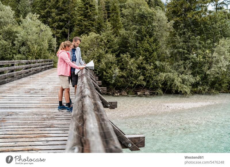 Young couple on a hiking trip reading map on wooden bridge, Vorderriss, Bavaria, Germany touristic tourists maps coat coats jackets go going walk hike relax