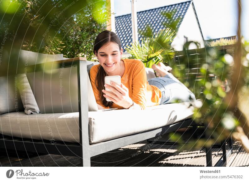 Young woman lying on couch on terrace using cell phone and laptop autonome autonomy independent funny having fun human human being human beings humans person
