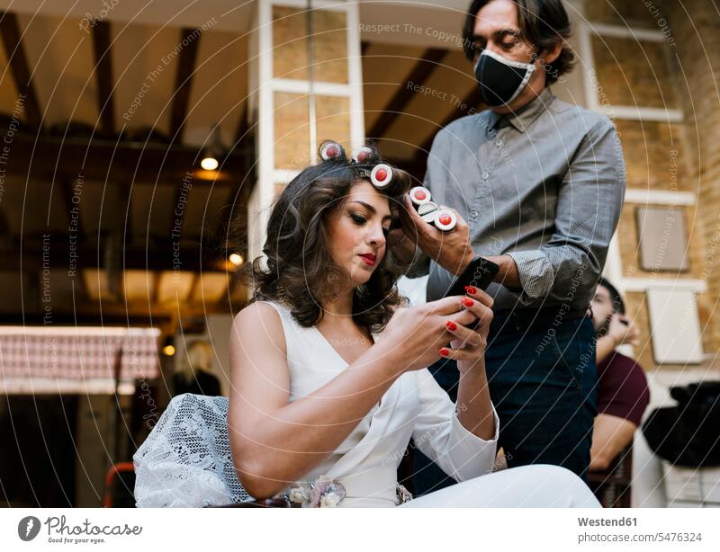 Male hairdresser doing hairstyle while bride using smart phone at salon during pandemic color image colour image indoors indoor shot indoor shots interior