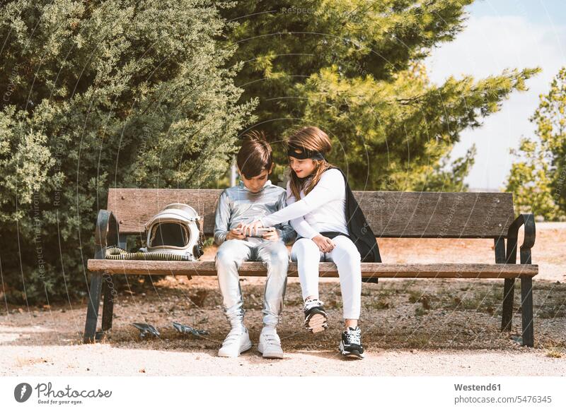 Two kids in astronaut and superhero costumes using mobile phone on park bench friends mate human human being human beings humans person persons braver bravers