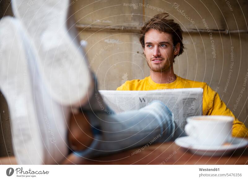 Portrait of young man with newspaper relaxing with feet up in a coffee shop newspapers shoes Tables read Seated sit drink relaxation free time leisure time