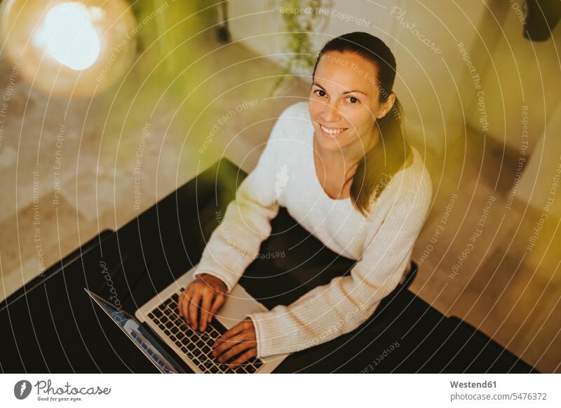 Smiling woman sitting with laptop at kitchen island color image colour image Connection connected connectivity Connections domestic kitchen kitchens