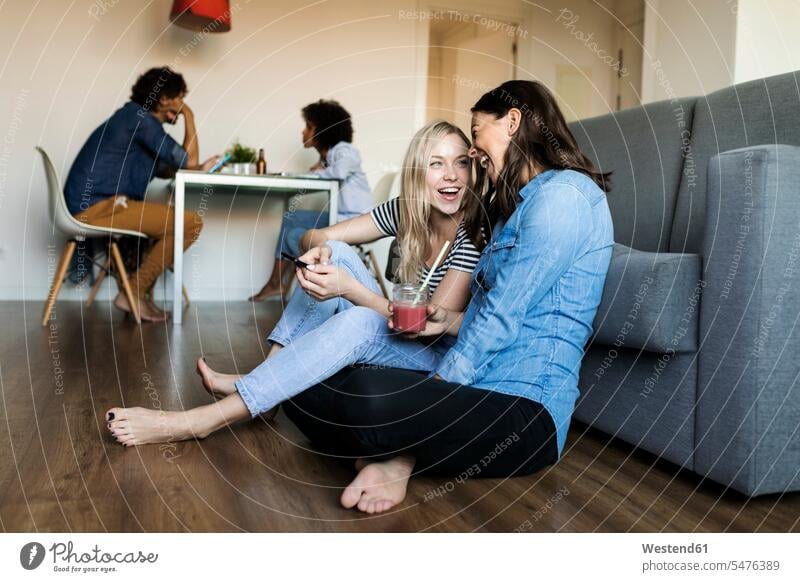 Two cheerful young women sitting on floor with cell phone and drink and friends in background female friends floors woman females gaiety Joyous glad