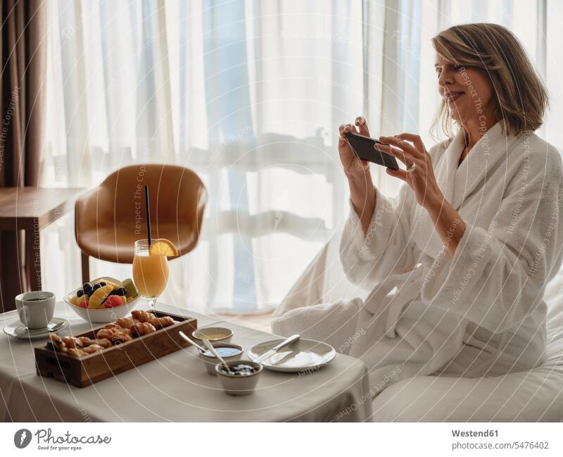 Smiling blond elderly woman photographing breakfast through smart phone while sitting on bed in hotel room color image colour image indoors indoor shot