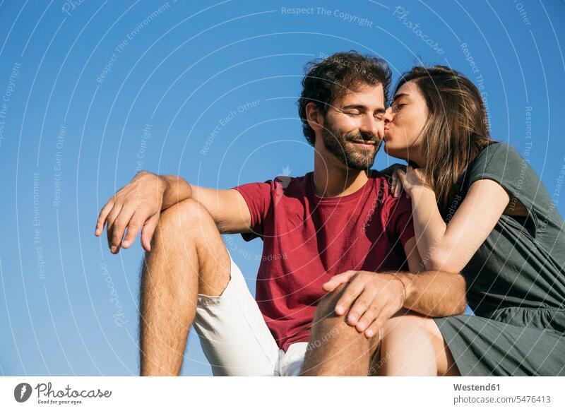 Young woman kissing man while sitting against clear sky color image colour image outdoors location shots outdoor shot outdoor shots day daylight shot
