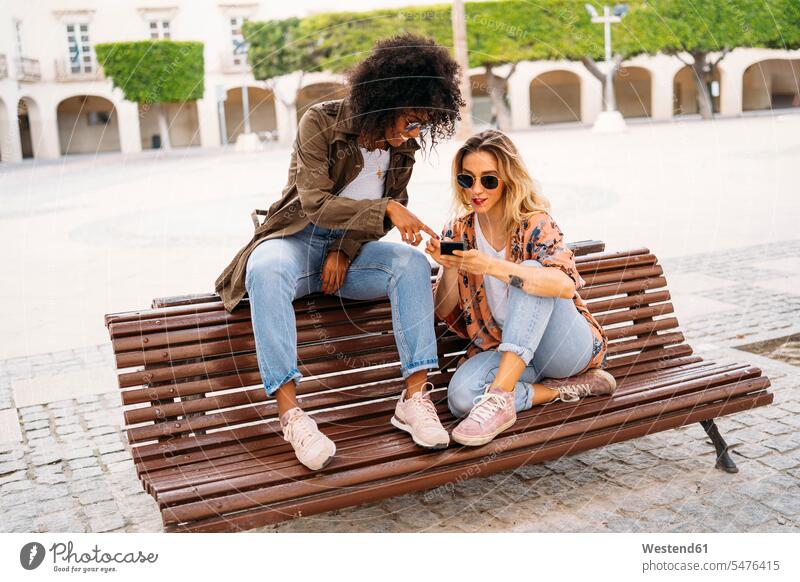 Multicultural happy women talking and using smartphone, sitting on bench benches telecommunication phones telephone telephones cell phone cell phones Cellphone