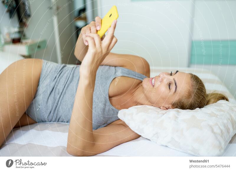Smiling woman text messaging through smart phone while lying on bed color image colour image Spain indoors indoor shot indoor shots interior interior view
