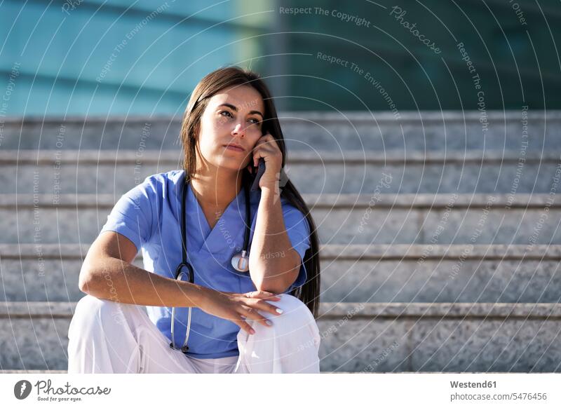 Young doctor talking on mobile phone while sitting on staircase against hospital color image colour image outdoors location shots outdoor shot outdoor shots day