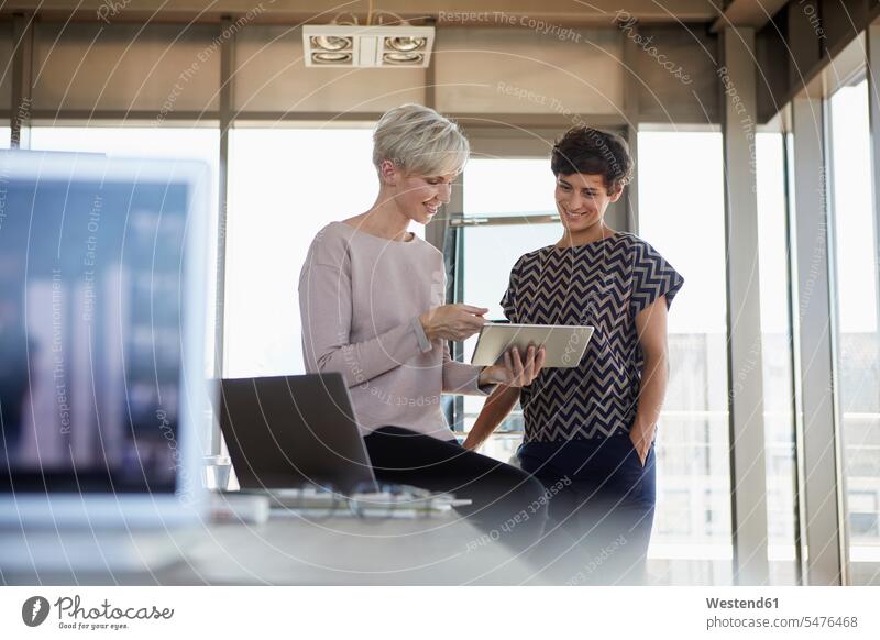 Two smiling businesswomen sharing tablet in office smile share working At Work offices office room office rooms businesswoman business woman business women