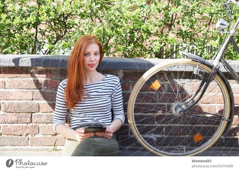 Portrait of redheaded woman with tablet and bicycle sitting on a wall females women red hair red hairs red-haired Seated walls digitizer Tablet Computer