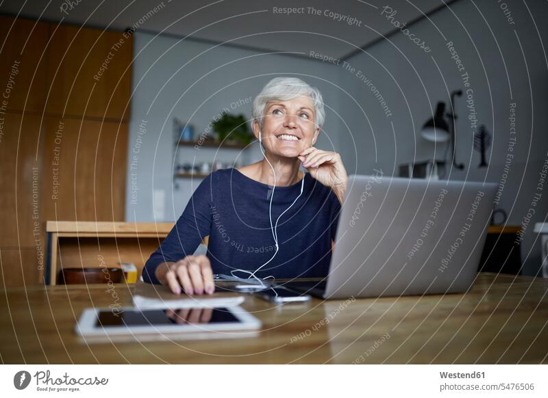 Active senior woman listening to music on smart phone and using laptop while looking away at home color image colour image indoors indoor shot indoor shots